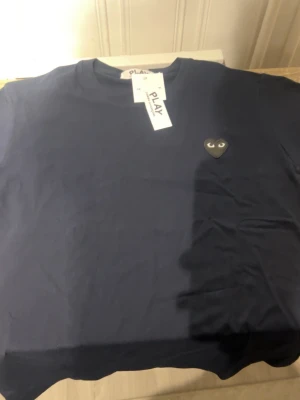 Mörkblå t-shirt Comme des Garçons Play - Mörkblå t-shirt från Comme des Garçons Play med det ikoniska hjärta-loggan i svart på bröstet. Klassisk rund halsringning och korta ärmar. Tillverkad i mjuk bomull för en skön känsla.