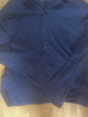 Mörkblå linne overshirt - En stilren mörkblå skjorta med långa ärmar och klassisk krage. Skjortan har knappar framtill och är tillverkad i ett mjukt bomullstyg. Perfekt för dig som gillar en enkel och clean look.
