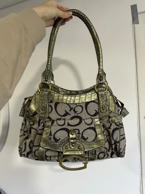 Vintage handväska Guess  - Snygg handväska i beige tyg med svart logomönster och detaljer i guldigt, krokopräglat material. Väskan har dubbla handtag, dekorativt spänne framtill och flera metalldetaljer. Perfekt accessoar för dig som vill sticka ut med en unik och trendig look.