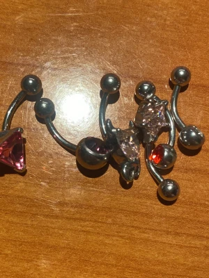 Smyckespaket med navelpiercingar - Flera navelpiercingar i silverfärgad metall med olika former av stenar i rosa, rött och klart.  Tjocklek är 1,6mm och längd är 10mm och en är 12 mm. Säljes alla blandade för 50kr