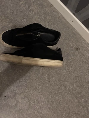 Arigato - Säljer ett par svarta sneakers med vit sula och dragkedja på ovansidan. Skorna har en låg profil och är tillverkade i ett material som ser ut som mocka eller sammet. Perfekta för en stilren och enkel look.