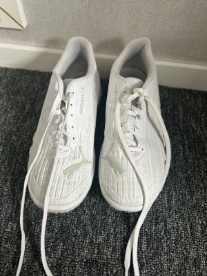 Vita Puma Adrenalite sneakers - Snygga vita sneakers från Puma, modell Adrenalite. Skorna har en låg silhuett, vita snören och diskret mönstrad ovandel i textil. Puma-loggan syns på tån och texten 'ADRENALITE' på plösen. Perfekta för en clean och sportig look.