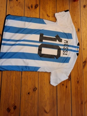Argentina Messi matchtröja blåvit - Säljer en klassisk Argentina matchtröja med blå och vita ränder, nummer 10 och Messi på ryggen. Tröjan har korta ärmar, svarta detaljer och klubbmärke framtill. Perfekt för fotbollsfans som vill stötta sitt lag.