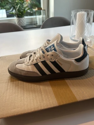 Adidas Samba vita och svarta sneakers - Klassiska Adidas Samba sneakers i vitt och svart med de ikoniska tre ränderna på sidan. Ovandelen är i läder med detaljer i mocka framtill och gummisula. Tungan har blå Adidas-logga och modellen har en låg profil som passar till många stilar.