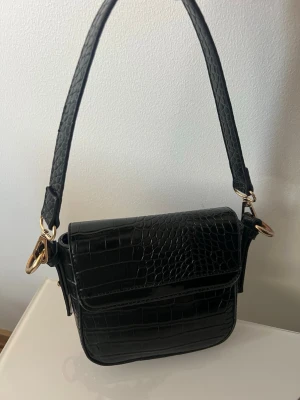 Svart axelremsväska i crocopräglat skinn - Stilren svart handväska i crocopräglat konstläder med bred axelrem och guldfärgade metalldetaljer. Väskan har en fyrkantig form och lockstängning. Perfekt för dig som vill ha en trendig accessoar med lyxig känsla.