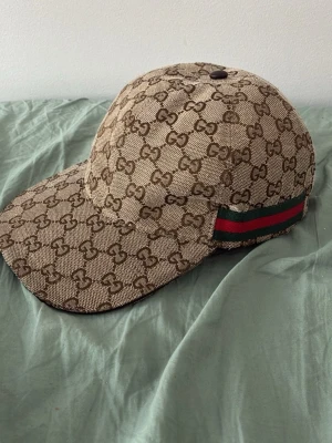 Beige GG-mönstrad keps från Gucci - Säljer en beige keps från Gucci med det klassiska GG-monogrammet över hela kepsen. På sidan finns en grön och röd rand som ger extra lyxig känsla. Kepsen har böjd skärm och är tillverkad i canvasmaterial. Perfekt för dig som vill ha en ikonisk och trendig accessoar.
