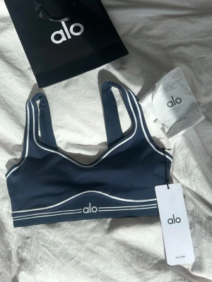 Mörkblå sport-bh från Alo - Helt oanvänd köpt för 3 dagar sen i Frankrikes egna Alo butik, Sport-bh i mörkblått med vita kontrastlinjer från Alo. Bh:n har breda axelband, rundad urringning och elastisk resår under bysten med Alo-logga framtill. Tillverkad i ett stretchigt och mjukt material som passar perfekt för träning och yoga.