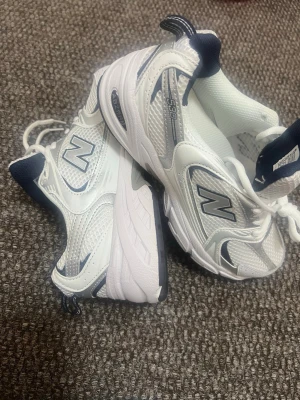Vita New Balance 530 sneakers - Snygga vita New Balance 530 sneakers med marinblå detaljer och meshpaneler för extra ventilation. Klassisk siluett med bekväm sula och tydlig N-logga på sidan. Perfekta för dig som gillar retrostil och sportig vibe. Obs det finns ingen shoebox!