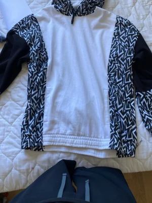Vit och svart hoodie från EA7 Armani - Säljer en snygg hoodie från EA7 Emporio Armani i vitt med svarta och vita grafiska mönster på ärmar och huva. Hoodien har dragsko i huvan och är tillverkad i mjukt material, perfekt för en chill och trendig look.