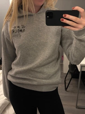 Grå hoodie med text och stjärna - Säljer en grå hoodie med svart text 'Je m'en amour' och en liten stjärna tryckt på bröstet. Hoodien har huva och ribbade muddar vid ärmslut och nederkant. Perfekt för en avslappnad och chill stil. SAKNAR SITT SNÖRE I HUVAN!!