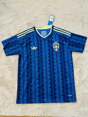 Blå Sverige fotbollströja Adidas - Säljer en blå fotbollströja från Adidas med Sveriges landslagsemblem på bröstet. Tröjan har ett mönster i olika blå nyanser, gula detaljer på axlarna och vit Adidas-logga. Tillverkad i lätt och ventilerande material, perfekt för träning eller match.