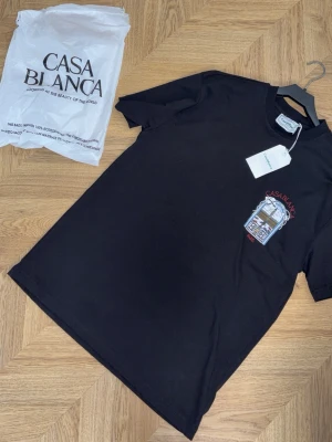 Svart t-shirt från Casablanca - Snygg svart t-shirt från Casablanca med tryck på bröstet som visar ett fönster och texten 'Casablanca Paris'. Klassisk rund hals och korta ärmar. Perfekt för dig som gillar stilrena plagg med en twist.