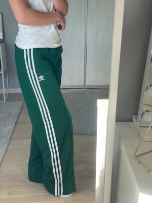 Gröna vida Adidasbyxor! - Säljer ett par gröna mjukisbyxor från Adidas med klassiska vita ränder längs benen och Adidas-logga på låret. Byxorna har en avslappnad passform och raka ben. Perfekta för chill dagar eller när du vill ha en sportig look.