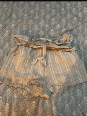 Beige randiga shorts med knytband - Säljer ett par ljusbeige shorts med vita ränder och knytband i midjan. Shortsen har hög midja, mjukt, luftigt tyg som känns perfekt för varma dagar. Snygga volangdetaljer upptill och avslappnad passform.
