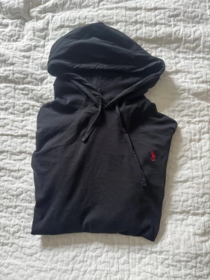 Ralph Lauren hoddie tunt material - Säljer en svart hoodie med huva och dragsnören. På bröstet finns en liten röd broderad logga. Hoodien har en klassisk passform och är tillverkad i mjukt material som känns skönt mot huden. Perfekt för en avslappnad och stilren look.