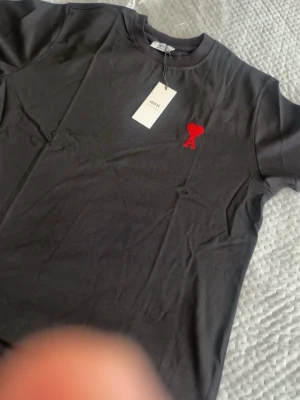 Svart t-shirt med rött hjärta Ami - Svart t-shirt från Ami med ett broderat rött hjärta och A på bröstet. Klassisk rund halsringning och korta ärmar. Tillverkad i mjuk bomull för en skön känsla och stilren look.