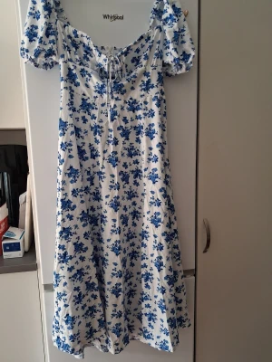 Vit-blå blommig långklänning - Superfin långklänning med vit botten och blått blommönster. Klänningen har puffärmar, snörning framtill och en lätt v-ringad urringning. Perfekt för sommarens alla tillfällen och ger en romantisk vibe.