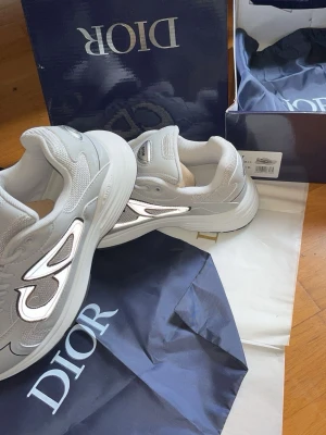 Grå Dior sneakers med reflekterande detaljer - Snygga grå sneakers från Dior med reflekterande detaljer på sidan och meshpaneler för extra andningsförmåga. Skorna har en chunky sula och klassisk snörning. Perfekt för dig som vill ha en exklusiv och trendig look.