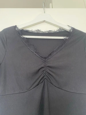 Svart långärmad topp med spets - Säljer en svart långärmad topp med ribbad struktur och v-ringning dekorerad med spets. Toppen har en snygg rynkning framtill som ger en feminin touch. Perfekt att styla med jeans eller kjol för en trendig look.