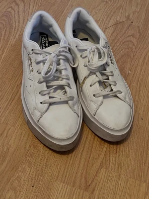 Vita Adidas sneakers med platåsula - Säljer ett par vita Adidas sneakers med platåsula och gulddetaljer vid loggan. Skorna har klassisk låg profil, vita skosnören och är tillverkade i slätt läder. Perfekta för dig som gillar stilrena och tidlösa sneakers med lite extra höjd.