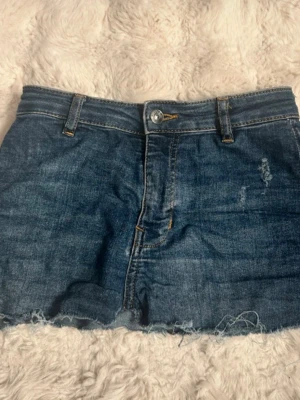 Blå jeanskjol från Divided - Säljer ett par klassiska blå jeanskjol från Divided. De har råa, fransiga kanter och normal passform. Kjolen har bälteshällor, knapp och dragkedja framtill samt slitna detaljer för en avslappnad look.