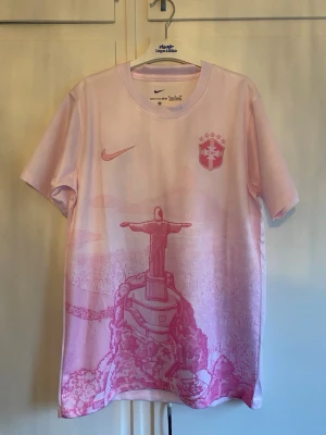 Rosa Nike fotbollströja Brasilien - Säljer en rosa fotbollströja från Nike med Brasilien-emblem och motiv av Kristusstatyn på framsidan. Tröjan har rund hals, korta ärmar och är tillverkad i ett lätt, ventilerande material som passar perfekt till träning eller match.