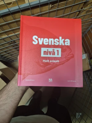 Svenska nivå 1 Helt enkelt - Lärobok i svenska för nybörjare på nivå 1. Boken har en röd och rosa framsida med vit och röd text samt en fiskillustration. Perfekt för dig som vill lära dig grunderna i svenska språket.