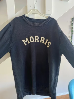 Mörkblå stickad tröja Morris - Mörkblå stickad tröja från Morris med rund halsringning och ribbade muddar vid ärmslut och nederkant. Tröjan har vit broderad MORRIS-text på bröstet och är tillverkad i ett mjukt stickat material. Perfekt för en avslappnad och stilren look.