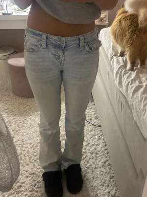 Ljusblå bootcut jeans från True Religion - Säljer ett par ljusblå bootcut jeans från True Religion med klassiska detaljer och broderade bakfickor. Jeansen har låg midja, fem fickor och är tillverkade i denim med en lätt tvättad look. Perfekta för en avslappnad och trendig stil.