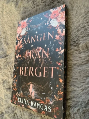 Sången från berget av Elina Kangas - En snygg pocketbok med titeln 'Sången från berget' av Elina Kangas. Omslaget är mörkt med rosa och vita blommor, stjärnor och en mystisk skogsmiljö. Boken har ett fantasifullt och magiskt tema, perfekt för dig som gillar äventyr och fantasy.