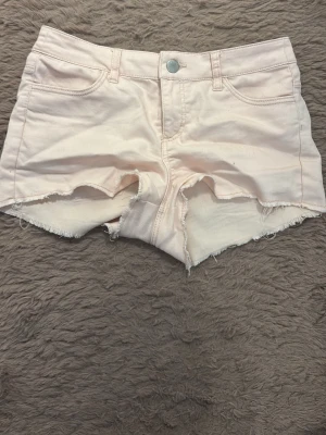 Ljusrosa jeansshorts med rå kant - Säljer ett par ljusrosa jeansshorts med fransig, rå kant nertill. Shortsen har fem fickor, bälteshällor och knappgylf. Perfekta för sommaren och ger en avslappnad vibe till din outfit.