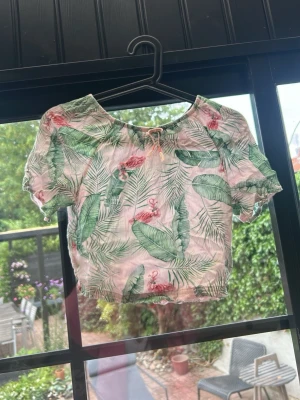 Grön-rosa croppad topp H&M x Karla - Säljer en croppad topp från H&M x Karla med tropiskt mönster av gröna blad och rosa flamingos. Toppen har korta puffärmar, resår i midjan och vid halsen samt en liten rosett framtill. Tunt, luftigt material perfekt för sommaren.