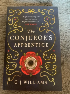 The Conjuror's Apprentice bok - En engelsk skönlitterär bok med svart omslag, dekorerad med guldfärgade slingor och en stor röd-vit blomma. Skriven av G J Williams och utgiven av Legend Press. Boken har ett mystiskt och historiskt tema med inslag av övernaturligt.