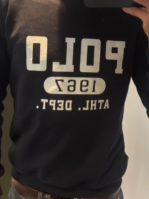 Mörkblå sweatshirt Polo 1967 - Mörkblå sweatshirt från Polo Ralph Lauren med trycket 'POLO 1967 ATHL. DEPT.' i vitt framtill. Klassisk rund halsringning och ribbade muddar vid ärmslut och nederkant. Perfekt för en avslappnad och sportig stil.
