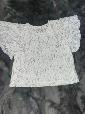 Vit spetsblus med puffärmar - Supersöt vit blus i spets med blommigt mönster och puffiga korta ärmar. Blusen har rund halsringning och är gjord i ett genombrutet spetsmaterial.