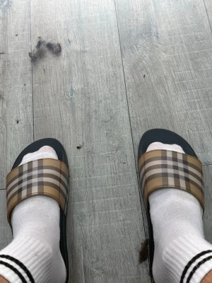Rutiga slides med beige och svart - Säljer ett par trendiga slides med klassiskt rutigt mönster i beige, svart och vitt. Tofflorna har en bred rem över foten och svart sula. Perfekta för chill dagar eller när du vill ha en avslappnad look.