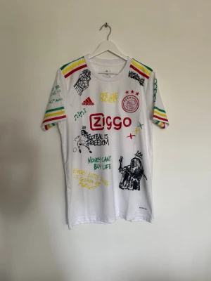Vit Bob Marley X Ajax  - Unik vit t-shirt med färgglada detaljer och tryck inspirerade av reggae och Bob Marley. T-shirten har korta ärmar med ränder i rött, gult och grönt samt flera grafiska tryck och texter både fram och bak. Perfekt för dig som gillar streetwear och statement-plagg.