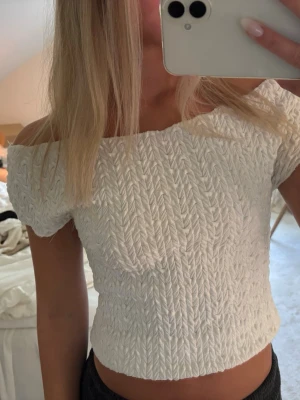 Vit offshoulder topp med struktur - Trendig vit offshoulder topp med korta ärmar och snygg struktur i tyget. Toppen är croppad och har en smickrande passform som sitter tajt mot kroppen. Perfekt att styla med både jeans och kjol för en cool look.
