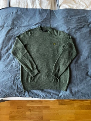 Grön stickad Lyle & Scott tröja - Mysig grön stickad tröja med rund hals och ribbade muddar. På bröstet sitter en liten gul broderad logga som detalj. Perfekt för lager på lager och enkel att matcha med jeans eller chinos. 