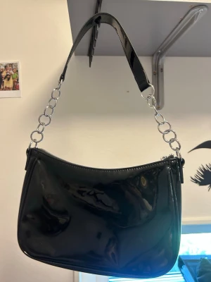 Svart axelväska i lack med kedjedetaljer - Snygg svart handväska i glansigt lackmaterial med silverfärgade kedjedetaljer på axelremmen. Väskan har en mjukt rundad form och minimalistisk design som ger en trendig look. Perfekt för dig som gillar statement-accessoarer.