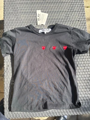 Svart t-shirt med röda hjärtan - Svart t-shirt från Comme des Garçons Play med tre små röda hjärtan broderade på bröstet. Klassisk rund hals och korta ärmar. Perfekt för dig som gillar stilrena plagg med en twist.