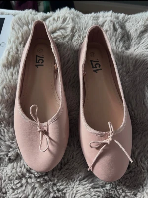 Ljusa rosa ballerinaskor med rosett - Säljer ett par ljusrosa ballerinaskor med rund tå och söt rosettdetalj framtill. Skorna har en enkel och klassisk design i ett mjukt material som känns bekvämt på foten. Perfekta för dig som gillar stilrena och feminina skor.