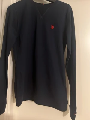 Mörkblå långärmad tröja - En stilren mörkblå långärmad tröja med rund halsringning och diskret röd broderad logga på bröstet. Tröjan har en klassisk passform och är tillverkad i ett mjukt material som passar perfekt till vardagslooken. 14-15