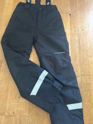 Svarta täckbyxor med reflexdetaljer - Svarta täckbyxor med hängslen och reflexdetaljer på benen. Byxorna har justerbar midja och är perfekta för kalla dagar utomhus. Märket cross sportwear syns på ena benet. Praktiska och snygga för vinterns alla äventyr.