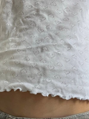 Vit ärmlös topp med hjärtmönster - Söt vit ärmlös topp med små hjärtan som mönster. Toppen har en ribbad struktur och vågig kant nertill för en extra detalj. Perfekt för varma dagar och enkel att matcha med olika stilar.