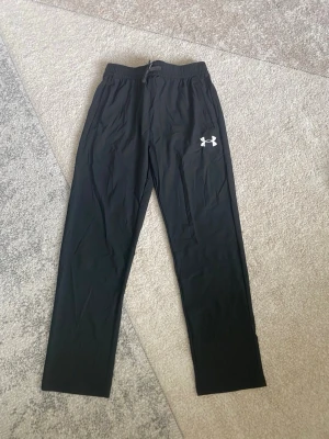 Under Armour byxor - Säljer ett par svarta träningsbyxor från Under Armour med resår i midjan och logga på vänster lår. Byxorna har raka ben och är tillverkade i ett lätt, snabbtorkande material som passar perfekt till gymmet eller löpning.
