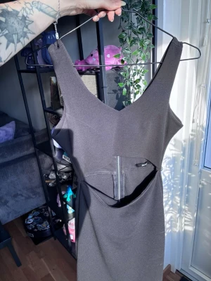 Svart klänning med cut-out och dragkedja - Snygg svart klänning med v-ringning, cut-out detalj i midjan och dragkedja bak. Klänningen är ärmlös och har en tight passform som framhäver figuren. Perfekt för dig som vill sticka ut med en edgy look.