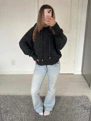 Mörkgrå stickad hoodie - Mysig mörkgrå stickad hoodie med luva och dragsko. Tröjan har en avslappnad passform, långa ärmar och ribbade muddar vid ärmslut och nederkant. Perfekt för dig som gillar en chill och enkel stil. Materialet ser ut att vara mjukt och värmande.