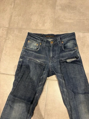 Blå jeans från Nudie Jeans Co - Säljer ett par klassiska blå jeans från Nudie Jeans Co med snygga slitningar och kontrastsömmar. Jeansen har fem fickor, knappgylf och detaljer i kopparfärg. Tillverkade i denim med en cool tvättad look.