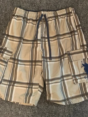 Beige rutiga cargoshorts med snörning - Snygga cargoshorts i beige med brunt och vitt rutmönster. Shortsen har snörning i midjan, flera fickor på sidorna och en avslappnad passform. Perfekta för sommardagar och chill häng.